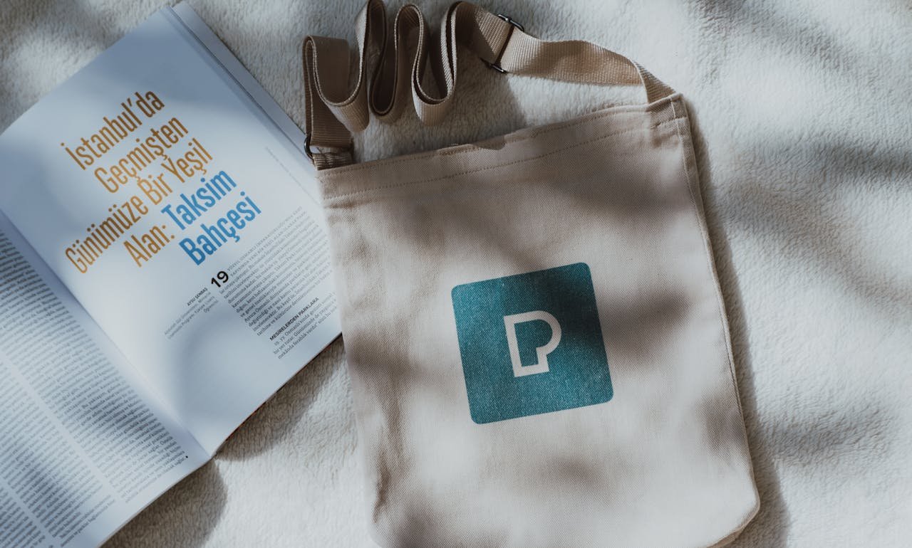 Top-down view of a tote bag with logo and magazine on soft light-colored fabric.